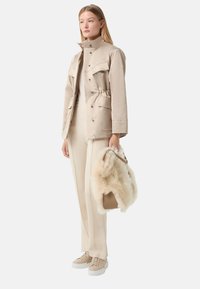 Veste imperméable beige avec des boutons pression et deux grandes poches avant, associée à un pantalon beige assorti et des baskets. Tient un sac doublé en fourrure.