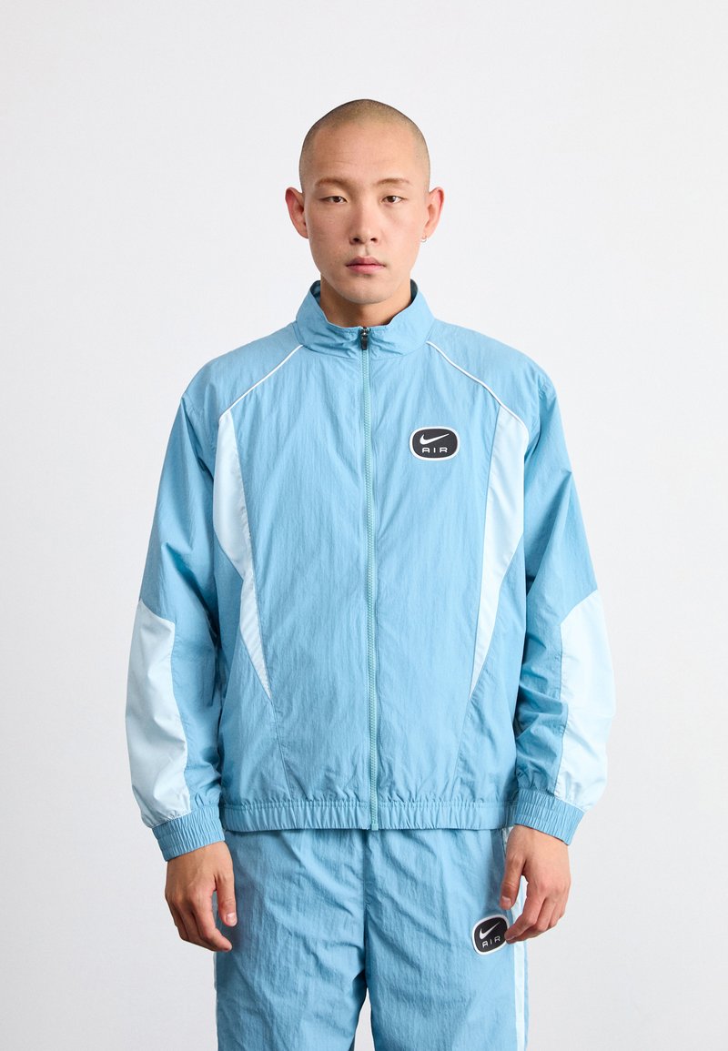 Ljusblå Nike-träningsjacka med hög krage, kontrasterande vita sidopaneler, elastiska ärmmuddar och en svart oval logotyp på bröstet.