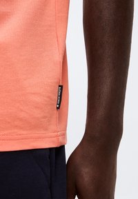 Κοντινό ενός σκοτεινόχρωμου χεριού δίπλα σε ένα ροδακινί πουκάμισο με ετικέτα Björn Borg και σκούρο μπλε παντελόνια.