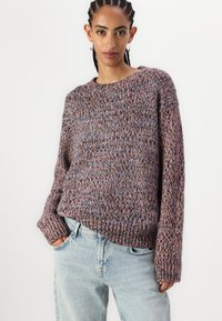 Strikkegenser i flerfargede fiber med rund hals, lange ermer, løs passform og ribbestrikkede mansjetter, parret med lyseblå jeans.