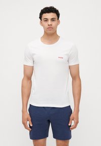 T-shirt en coton blanc avec col rond et manches courtes ; présente un logo rouge "HUGO" sur la poitrine gauche. Associé à un short bleu.