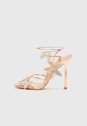 Rozā zelta krāsas augstpapēžu sandales ar tauriņa motīviem, izceļoties ar smalkiem siksniņām un dimanta imitācijas detaļām. Plakana zole ar kvadrātveida papēdi.