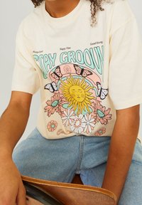 Krémové bavlněné tričko s barevným slunečním a květinovým motivem, s motýly a textem "STAY GROOVY." Uvolněný střih a krátké rukávy.
