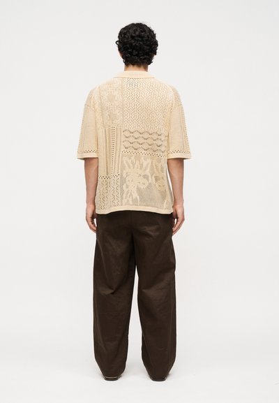 Άνδρας με σγουρά μαλλιά που φοράει μπεζ πλεκτό πουκάμισο με κοντά μανίκια και σχέδιο και χαλαρό σκούρο καφέ παντελόνι, στέκεται με την πλάτη προς ένα απλό φόντο.