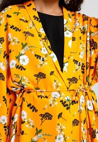 Floral kimono in vibrant orange with white and black flowers. Features a V-neck, wide sleeves, and a tie belt at the waist. Smooth texture.