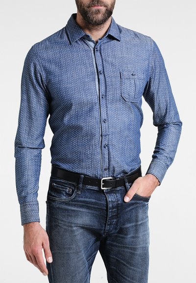 Denimskjorta med knappar och ett litet vitt prickmönster, långa ärmar och en bröstficka. Bärs tillsammans med mörka denimjeans och ett svart bälte.