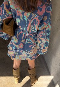 Άτομο που φοράει ζώνη, πολύχρωμο φόρεμα με ανθισμένο μοτίβο και μακριά μανίκια, μαζί με μπεζ σουέτ μπότες μέχρι το γόνατο, στέκεται σε τσιμεντένιο δάπεδο.