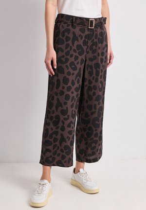 Personne portant un pantalon marron à jambes larges avec un imprimé léopard noir et une ceinture, assorti à des baskets blanches et un haut blanc.