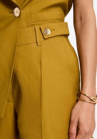 Pantalones mostaza con corte entallado, que presentan un bolsillo lateral, un cinturón decorativo con un botón metálico y una tela texturizada.