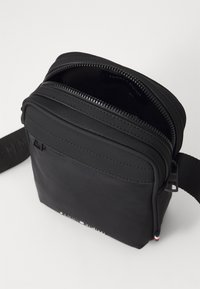 Svart nylon crossover-veske med glidelåslukking, rektangulær form, justerbar stropp og subtil merkevare på fronten.