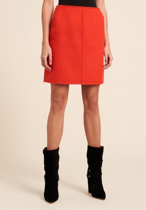 Mini-jupe en laine rouge avec une coupe droite, détail de couture à l'avant et poches latérales, associée à des bottes de cheville en suède noires.
