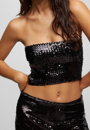 Frau trägt ein trägerloses schwarzes Pailletten-Crop-Top und passenden Rock, steht mit den Händen in der Hüfte, langes, welliges Haar über die Schulter gelegt.