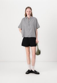 Raidallinen, lyhythihainen paita mustavalkoisena, napitettu, yhdistetty mustiin shortseihin. Asusteina pieni vihreä laukku ja mustat loaferit.