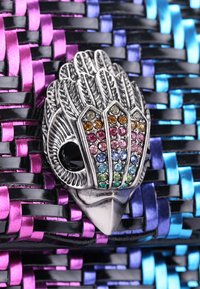 Srebrzysta broszka w kształcie ptaka z kolorowymi cyrkoniami na teksturowanym tle z plecionego czarnego, fioletowego i niebieskiego materiału.