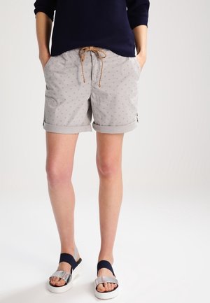 Persoon draagt lichtgrijze korte broek met patroon en opgerolde boorden, donkerblauwe top en zilveren sandalen met zwarte banden, staand tegen een effen achtergrond.