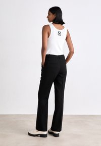 Femeie stând cu mâinile în buzunare, purtând un maiou alb fără mâneci cu logo pe spate, pantaloni negri și pantofi de culoare crem, cu spatele spre privitor.