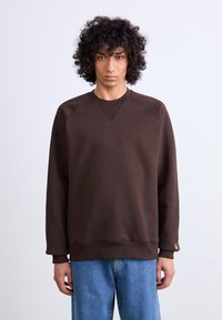 Brown sweatshirt with a crew neck, raglan sleeves, and ribbed cuffs. Notable detail includes a small logo tag on the left sleeve.