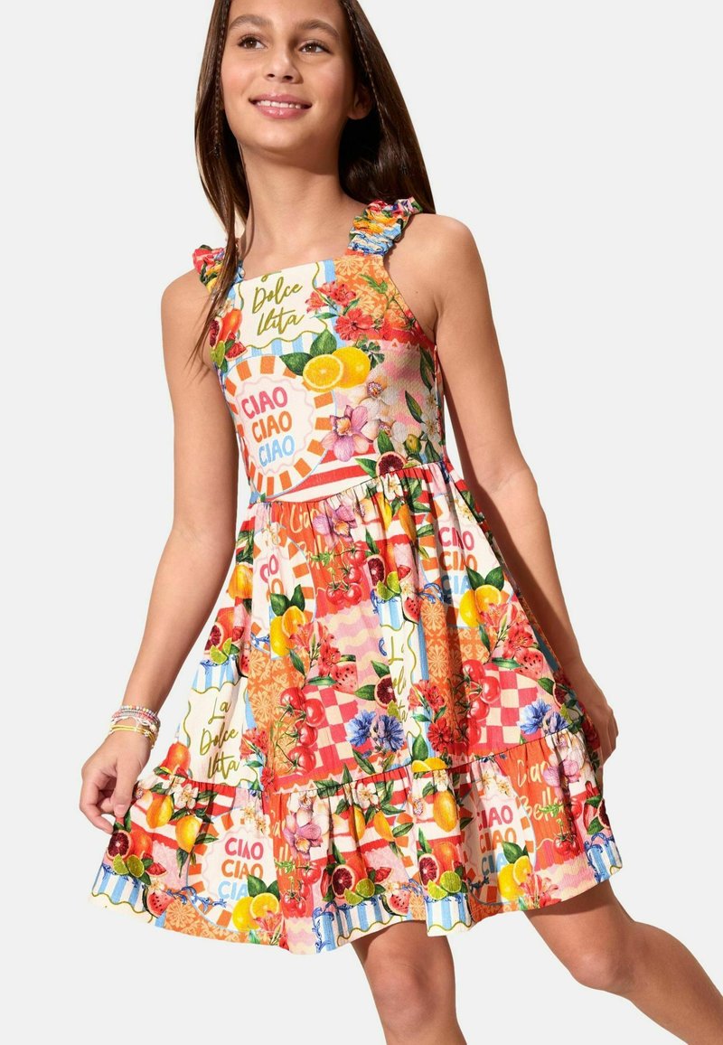 Niña con un vestido sin mangas colorido con tirantes fruncidos y un estampado de frutas, flores y palabras italianas como "ciao" y "Dolce Vita."