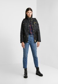 Black waterproof jacket with gold hardware, multiple pockets, and a high collar. Worn over a plaid shirt and paired with blue jeans and black boots.
