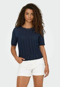 Navyblauwe gebreide top met korte pofmouwen, voorzien van een geribd patroon en een golvende zoom. Gecombineerd met witte opgerolde shorts.