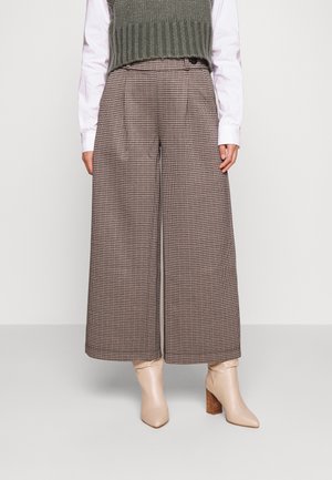 Woman wearing wide-leg brown checkered pants, white shirt, green knit vest, and beige heeled ankle boots standing on a plain background.