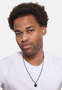 Zwarte halsketting met ronde hanger op een model in een wit T-shirt. Het model heeft donker, krullend haar en een neutrale uitdrukking.