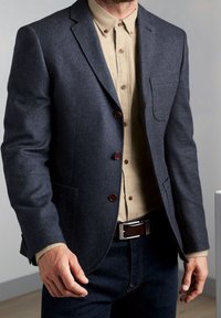 Námořnický sako ve vzoru herringbone s třemi hnědými knoflíky, nošeno přes béžovou košili, v kombinaci s tmavými džíny a hnědým páskem. Mezi detaily je také náprsní kapsa.