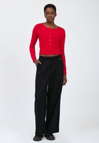 Cardigan rosso lavorato a maglia con maniche lunghe e chiusura a bottoni, abbinato a pantaloni neri a vita alta con gamba larga e scarpe nere con suola spessa.