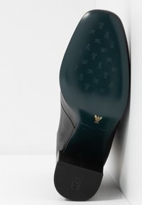 Chaussure en cuir noir avec une semelle texturée affichant un motif de papillon. Présente un logo accent doré près du talon. Fini lisse, forme arrondie.