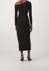 Černé midi šaty s dlouhým rukávem a jedním ramínkem, s přiléhavou siluetou, hladkou texturou a délkou do poloviny lýtek. Nosené s černými vysokými podpatky.
