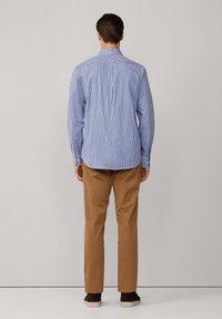Man in een blauw geruite overhemd met lange mouwen en een bruine broek, staand met zijn gezicht naar een lichtgrijze muur met ontspannen handen langs zijn zij.
