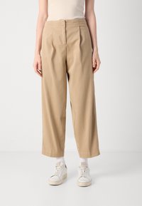 Beige wijde broek met geplooid voorkantontwerp, gemaakt van een lichtgewicht stof. Gecombineerd met witte sneakers en zichtbare crew-sokken.