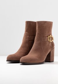 Bottines en daim marron avec un talon épais. Présente une sangle avec une boucle dorée sur le côté. Texture lisse, bout rond.