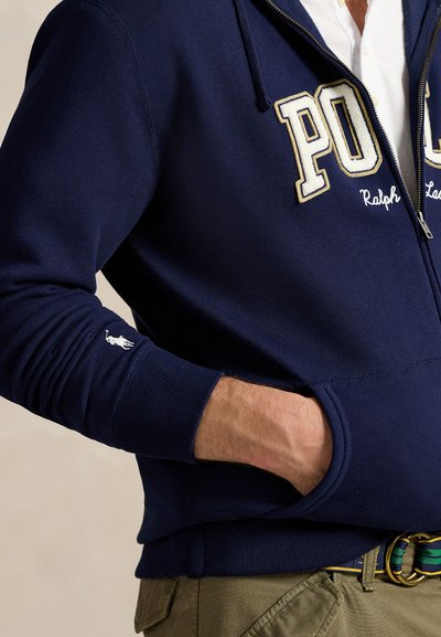 Mörkblå zip-up hoodie med vit och guld "Polo" bokstavering, ribbade manschetter och sidofickor. Har en liten vit hästlogotyp på ärmen.