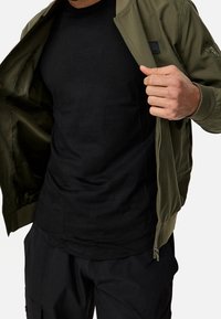 Homme portant un t-shirt noir à col rond et une veste bomber vert olive, tenant la veste ouverte pour dévoiler le t-shirt.