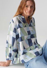 Blusa morbida e ampia con un motivo geometrico in blu navy, azzurro, verde e bianco. Tessuto morbido con scollo a V e maniche con polsini.