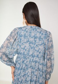 Abito floreale blu con maniche lunghe a sbuffo trasparenti e vita arricciata. Motivo floreale in tonalità chiare su un tessuto texturizzato.