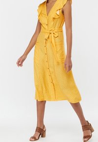 Žluté šaty s puntíky, ruffled rukávy, zapínání na knoflíky vpředu a zavazovací pásek v pase. Hnědé sandály na podpatku dokončují outfit.