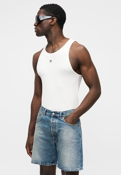 Biely rebrovaný tielkový top s čiernym logom, v kombinácii so svetlo modrými rifľovými šortkami s oteraným lemom a bočnými vreckami. Modelka má na sebe slnečné okuliare.