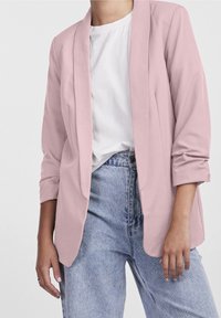 Světle růžový blejzr s volným střihem, otevřeným předním dílem a ohrnutými rukávy, zkombinovaný s bílým tričkem a světle modrými džínami s vysokým pasem.
