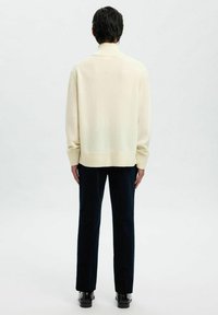 Cremfarbener Pullover mit hohem geripptem Kragen und langen Ärmeln, kombiniert mit dunklen Hosen und schwarzen Schuhen, von hinten betrachtet.