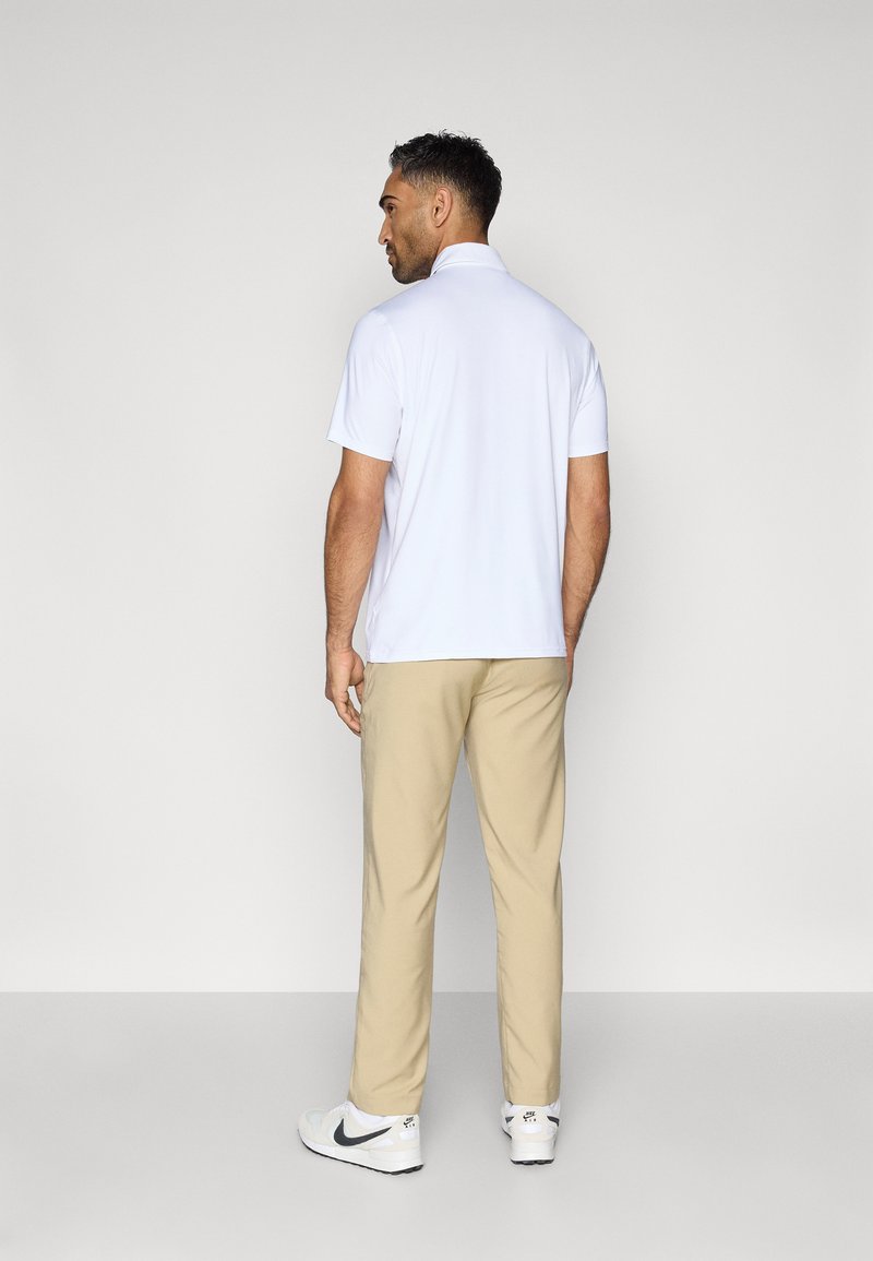 Polo shirt blanc à manches courtes, pantalon beige et sneakers blancs avec des accents noirs. La vue est prise par l'arrière, mettant en avant une coupe décontractée.