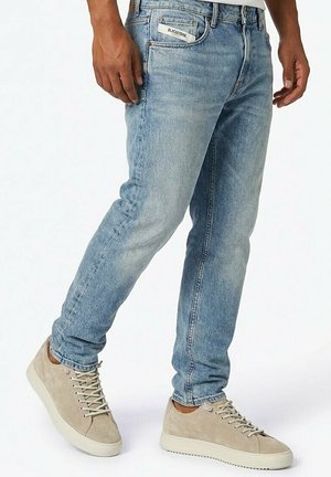 Man die lichtblauwe slim-fit jeans draagt met een wit label in de zak en beige suède sneakers, staand tegen een witte achtergrond.