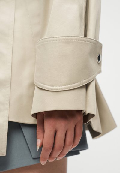 Χέρι με μακριά νύχια κάτω από μανίκι μπεζ σακακιού με δερμάτινο φιόγκο με κουμπί, που ακουμπάει κοντά στη γαλάζια φόδρα φούστας πάνω σε λευκό φόντο.