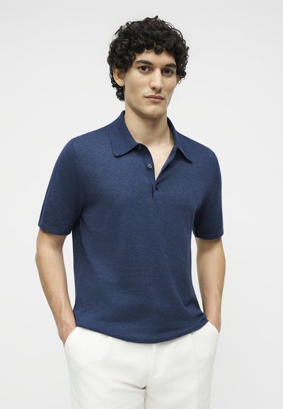 Νέος άνδρας με σγουρά μαλλιά φοράει μπλε πόλο με κοντά μανίκια και λευκό παντελόνι, στέκεται με τα χέρια στις τσέπες μπροστά σε απλό φόντο.