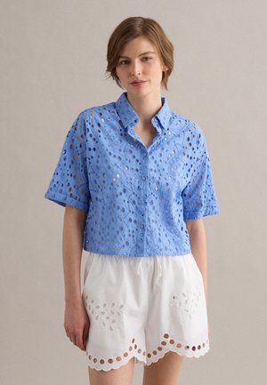 Mujer con camisa azul de manga corta con ojales y botones y pantalones cortos blancos con dobladillo ondulado y detalles bordados, de pie frente a un fondo liso.