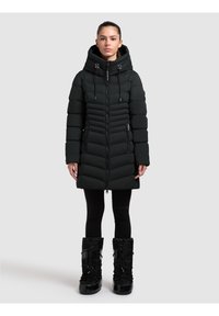 Čierny nadýchaný kabát s vysokým golierom, so zipsom vpredu, bočnými vreckami a prešívaným dizajnom. Kombinovaný s čiernymi snehovými topánkami.