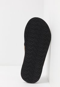 Semelle de chaussure en caoutchouc noir avec un motif en zigzag texturé, présentant un côté partiel en cuir brun, inclinée contre un fond blanc.