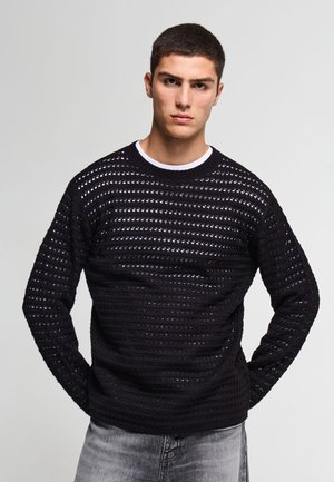 Giovane uomo con capelli corti e scuri che indossa un maglione nero a maglia aperta sopra una camicia bianca e jeans grigi sbiaditi, in piedi con le mani dietro la schiena.