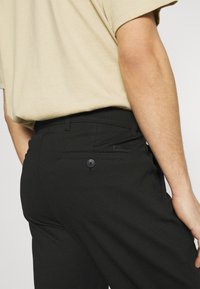 Schwarze Hose mit taillierter Passform, ausgestattet mit einer Gesäßtasche mit Knopfverschluss und einer glatten, eleganten Textur. Kombiniert mit einem beigen Hemd.